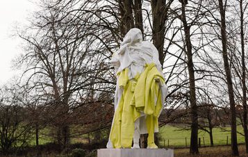 An abstract sculpture of a humanoid figure covered in draped fabric, displayed outdoors surrounded by trees.