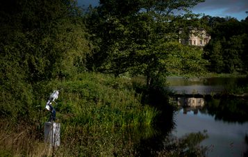 A female figure wearing jeans standing on a white plinth at the edge of a lake, surrounded by foliage. Bretton Hall is in the distance