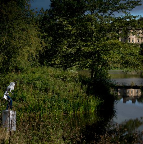 A female figure wearing jeans standing on a white plinth at the edge of a lake, surrounded by foliage. Bretton Hall is in the distance