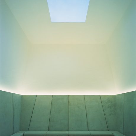 A room with concrete bench seating and a square hole cut into the ceiling revealing the sky