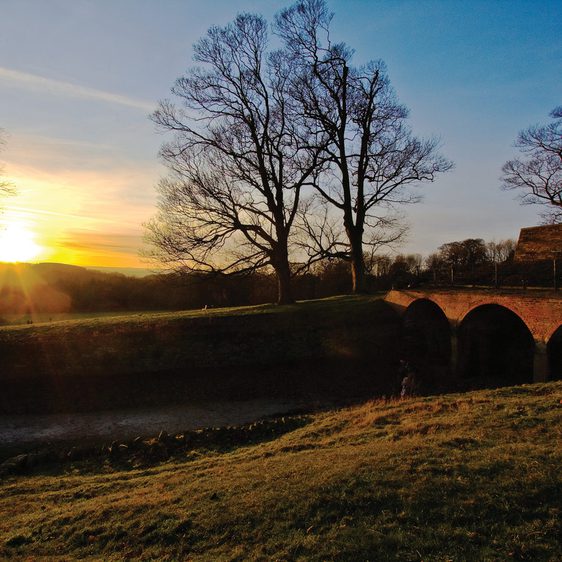 The sun rising over a brick deer shelter, surrounded by trees.