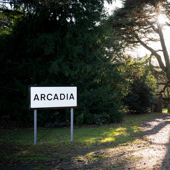 A white road sign with ARCADIA in black letters, outdoors near to some trees