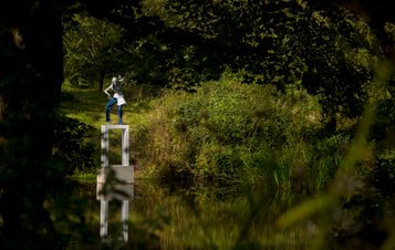 A female figure wearing jeans standing on a white plinth at the edge of a lake, surrounded by foliage