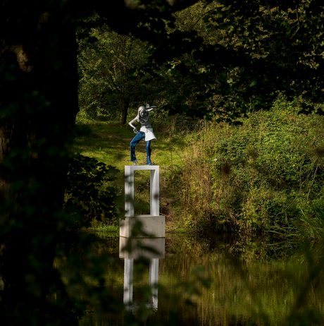A female figure wearing jeans standing on a white plinth at the edge of a lake, surrounded by foliage
