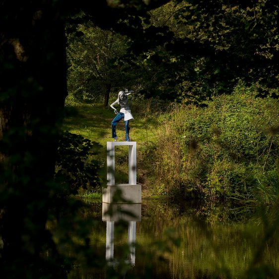 A female figure wearing jeans standing on a white plinth at the edge of a lake, surrounded by foliage