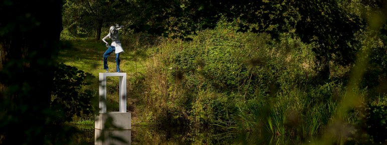 A female figure wearing jeans standing on a white plinth at the edge of a lake, surrounded by foliage