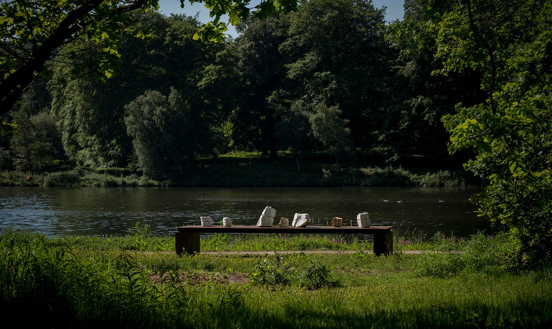A bronze table structure with white marble pieces wrapped in wire on top. The lake is in the background.