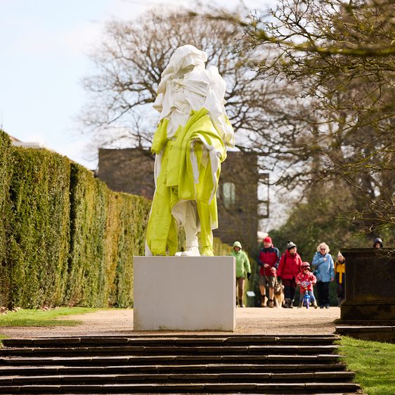 A group of people in colourful coats walking towards a yellow and white sculpture of a figure draped in clothing