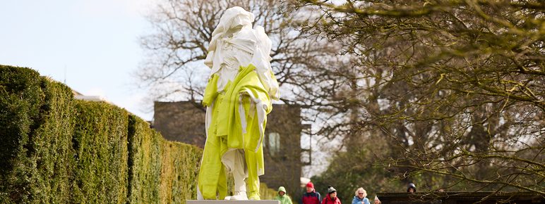 A group of people in colourful coats walking towards a yellow and white sculpture of a figure draped in clothing
