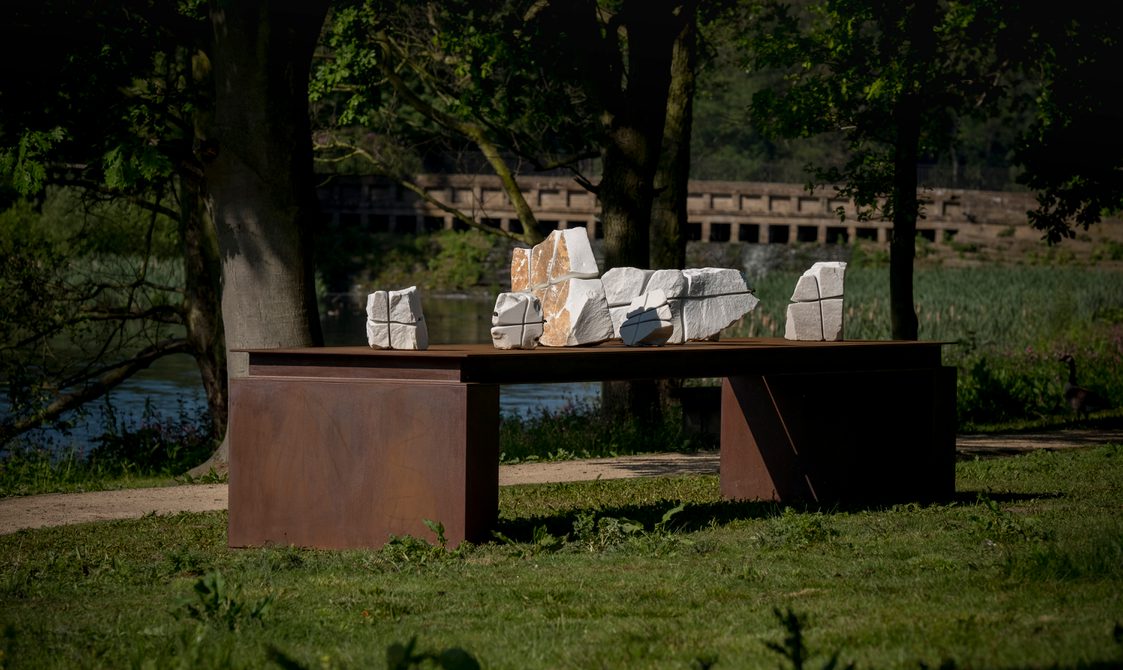 A bronze table structure with white marble pieces wrapped in wire on top. The cascade bridge is in the background.