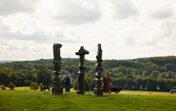 Dog walkers looking at three tall thin bronze sculptures looking out over the landscape