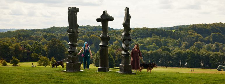 Dog walkers looking at three tall thin bronze sculptures looking out over the landscape