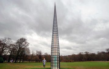 A person walking towards a tall pointed tower, with glass panels