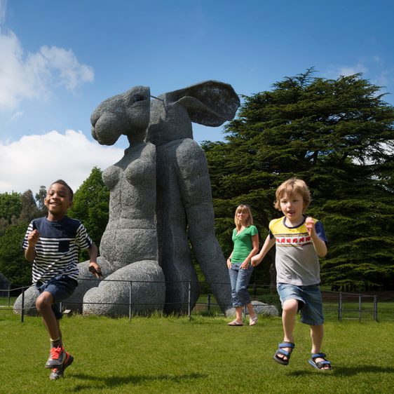 A group of children running in front of a large sculpture of a female body with a rabbit head.