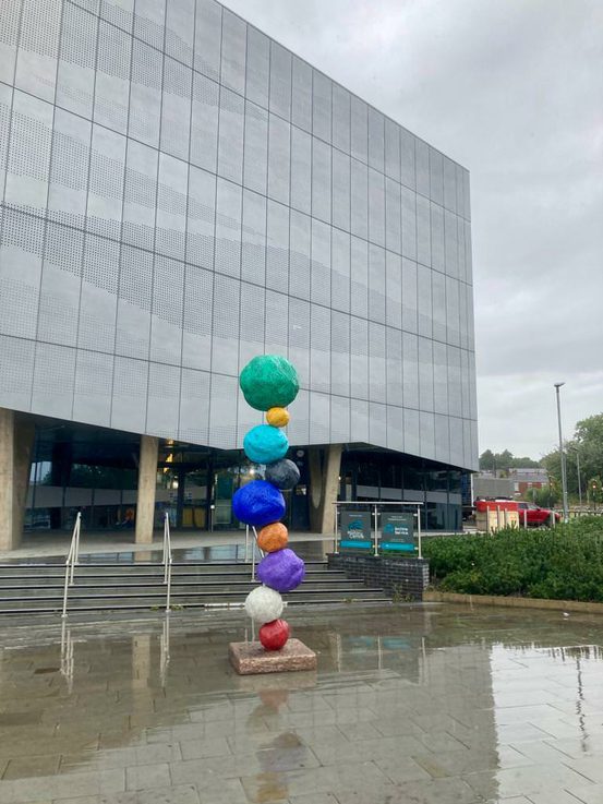 A brightly coloured stack of shapes in front of a grey building