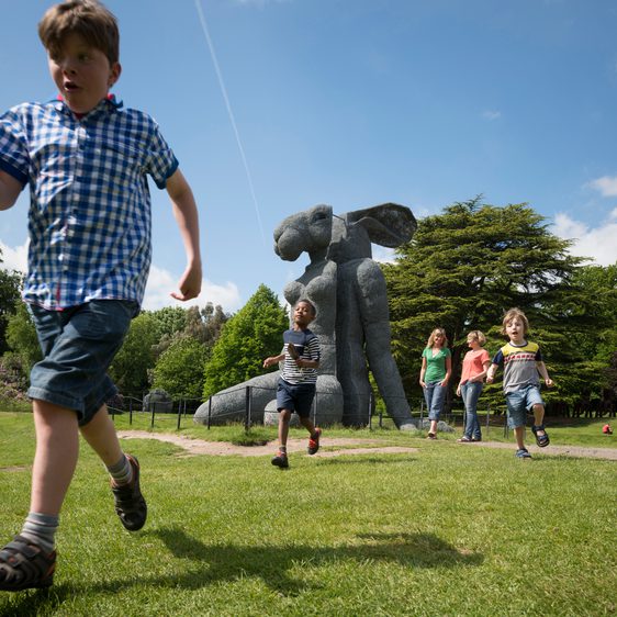 A group of children running in front of a large sculpture of a female body with a rabbit head.