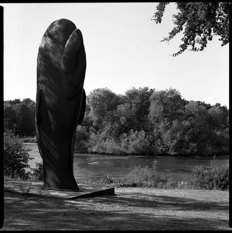 A black and white photo of a large abstract sculpture displayed near to a lake, surrounded by trees and foliage
