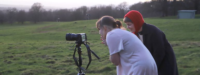 Two women, one wearing a pink night gown and one wearing a red knitted hood, looking into a camera outdoors.