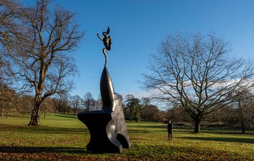 A man looking at a sculpture of a hare balanced on the tip of an oversized anvil, displayed outdoors.