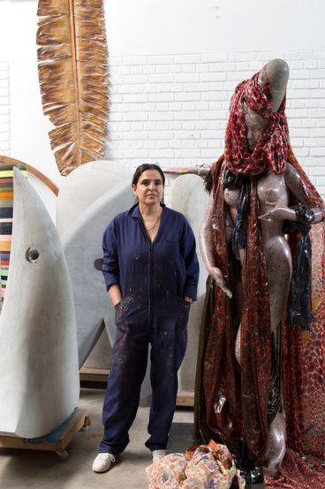 A South Asian woman wearing black overalls, standing between figurative sculptures in her studio.