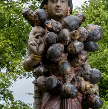 A large, intricately detailed sculpture of a woman holding multiple small figures, set against a backdrop of lush green trees. The woman has a serene expression, and the figures appear to be clinging to her.