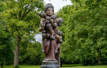 A large, intricately detailed sculpture of a woman holding multiple small figures, set against a backdrop of lush green trees. The woman has a serene expression, and the figures appear to be clinging to her.
