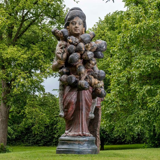 A large, intricately detailed sculpture of a woman holding multiple small figures, set against a backdrop of lush green trees. The woman has a serene expression, and the figures appear to be clinging to her.