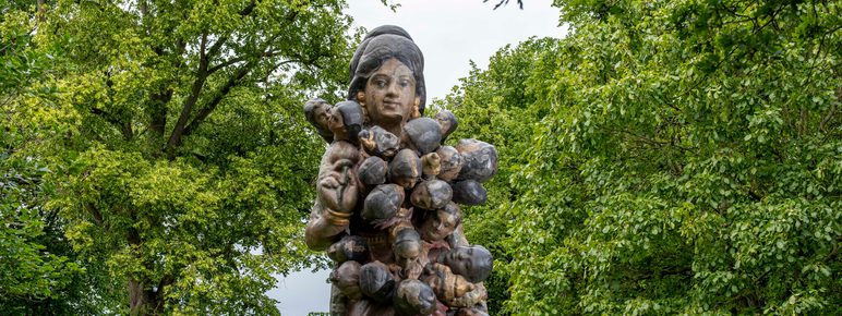 A large, intricately detailed sculpture of a woman holding multiple small figures, set against a backdrop of lush green trees. The woman has a serene expression, and the figures appear to be clinging to her.