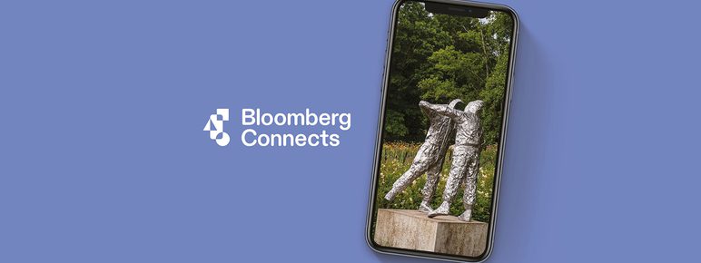 An image of a mobile phone displaying a photograph of a sculpture depicting two embracing figures. The image has a pale blue background with white text: Bloomberg Connects