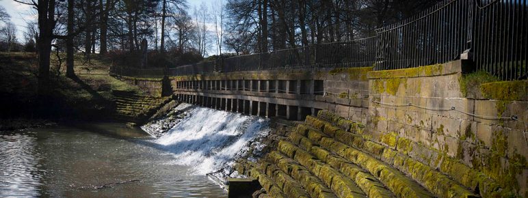 A stone weir with a gentle waterfall flows into a calm river. The surrounding area features moss-covered steps leading to the water, framed by trees and a wrought iron fence under a clear blue sky.