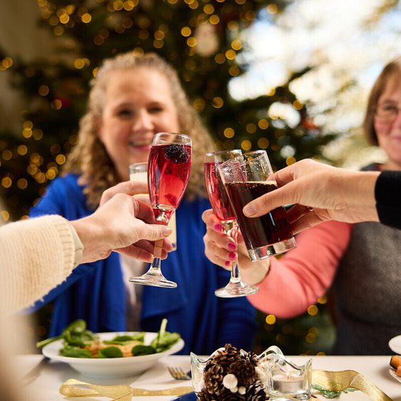A festive gathering featuring three people toasting with drinks at a table adorned with food. In the background, a decorated Christmas tree glimmers with lights.