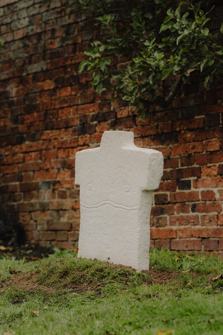 A white stone sculpture resembling a human torso stands on a patch of grass against a brick wall.
