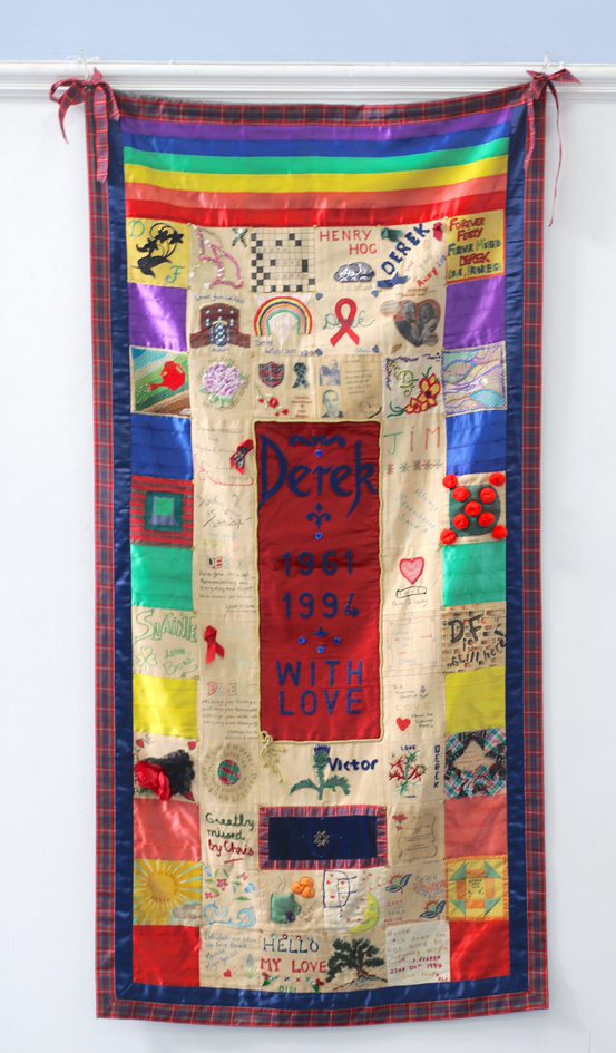 An embroidered textile panel featuring colourful squares and text memorialising Derek, who died from AIDS.