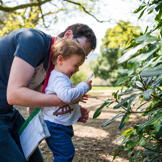 A man and a young child are exploring a garden. The man leans over the child, who is excitedly looking at a bush through a magnifying glass. Both are engaged in the activity, with the child wearing a playful expression. Sunlight filters through the trees in the background.