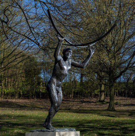 A bronze sculpture of a slender figure holding a large curved object above their head, set in a grassy area surrounded by trees. The sky is clear and blue, creating a serene atmosphere in the park.