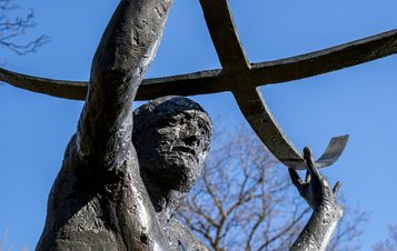 A close-up of a bronze statue depicting a muscular figure holding a large, curved crossbar above his head. The background features a clear blue sky and bare trees, suggesting a serene outdoor setting. The statue conveys strength and determination.