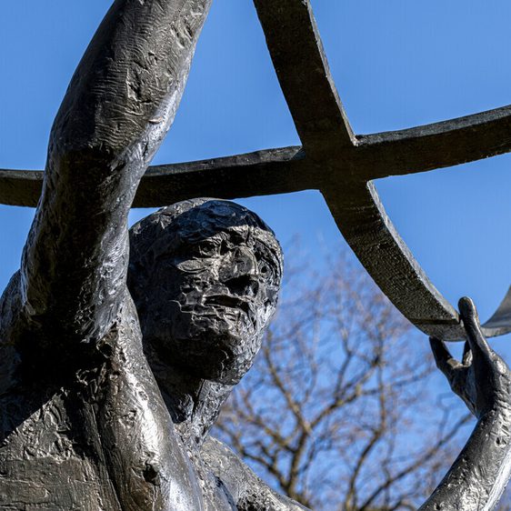 A close-up of a bronze statue depicting a muscular figure holding a large, curved crossbar above his head. The background features a clear blue sky and bare trees, suggesting a serene outdoor setting. The statue conveys strength and determination.