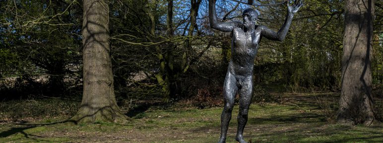 A bronze sculpture of a slender figure holding a large curved object above their head, set in a grassy area surrounded by trees. The sky is clear and blue, creating a serene atmosphere in the park.