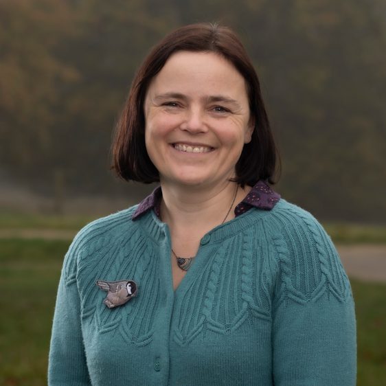 Women with short brown hair smiles, she is wearing a green cardigan with a bird broach