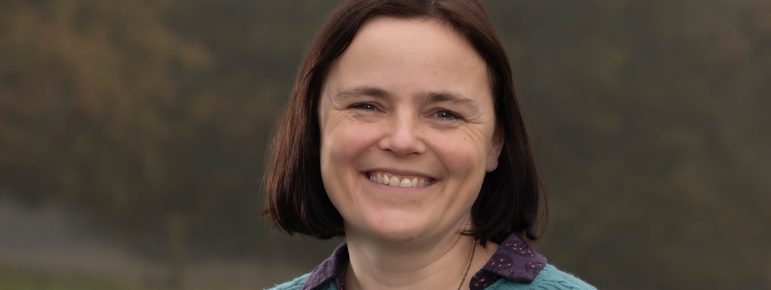 Women with short brown hair smiles, she is wearing a green cardigan with a bird broach