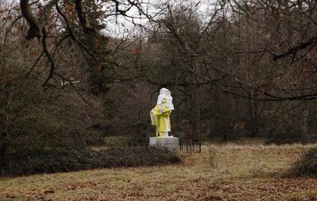 An abstract sculpture of a humanoid figure covered in draped fabric, displayed outdoors surrounded by trees.