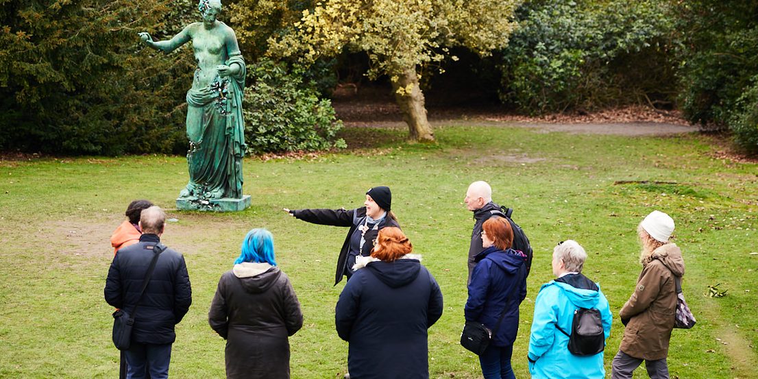 A group of people and a guide looking at a bronze sculpture of a woman outdoors