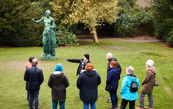 A group of people and a guide looking at a bronze sculpture of a woman outdoors