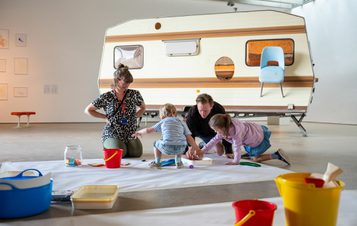 A group of people drawing on a huge piece of paper in front of a caravan artwork in the gallery