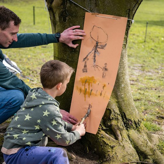 An adult and a child drawing together onto a large piece of paper leaning against a tree outdoors.