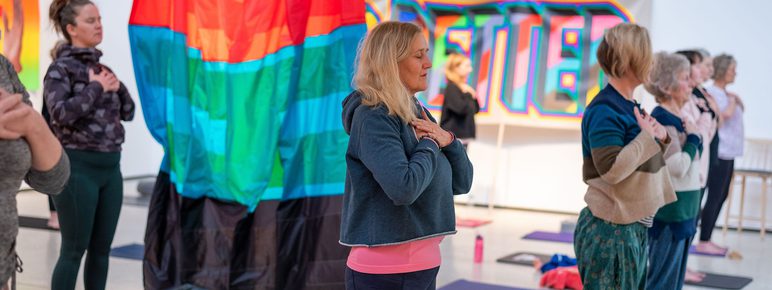A group of people practicing yoga inside a gallery space, with colourful fabric hangings and paintings.