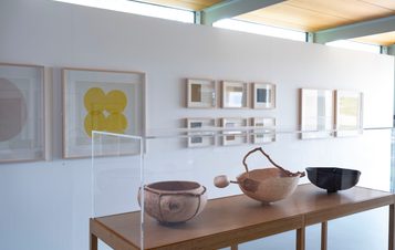 A white gallery space with framed prints on the walls and carved wooden vessels on a table in the centre