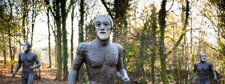 Three nude male bronze figures with white painted faces, displayed outdoors.