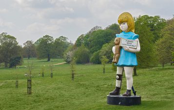 Sculpture of a young girl holding a teddy bear and a charity box, among saplings in YSP parkland.