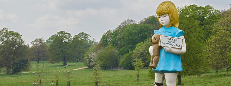Sculpture of a young girl holding a teddy bear and a charity box, among saplings in YSP parkland.